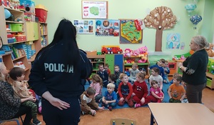 Sala lekcyjna. Policjantka stoi przed przedszkolakami, którzy siedzą na podłodze. .Policjantka zwrócona jest plecami do zdjęcia. W tle widać półki z zabawkami.