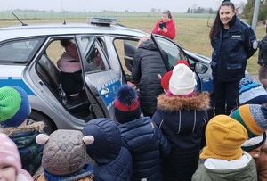 Teren przy szkole. Dzieci oglądają radiowóz policyjny, cześć z nich siedzi w środku pojazdu. Pośród nich stoi policjantka.