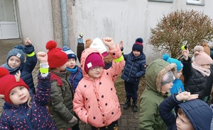 Dzieci ubrane w kurtki stoją przed budynkiem. Na podnoszonych do góry rękach załączone mają otrzymane od policjantek opaski odblaskowe.